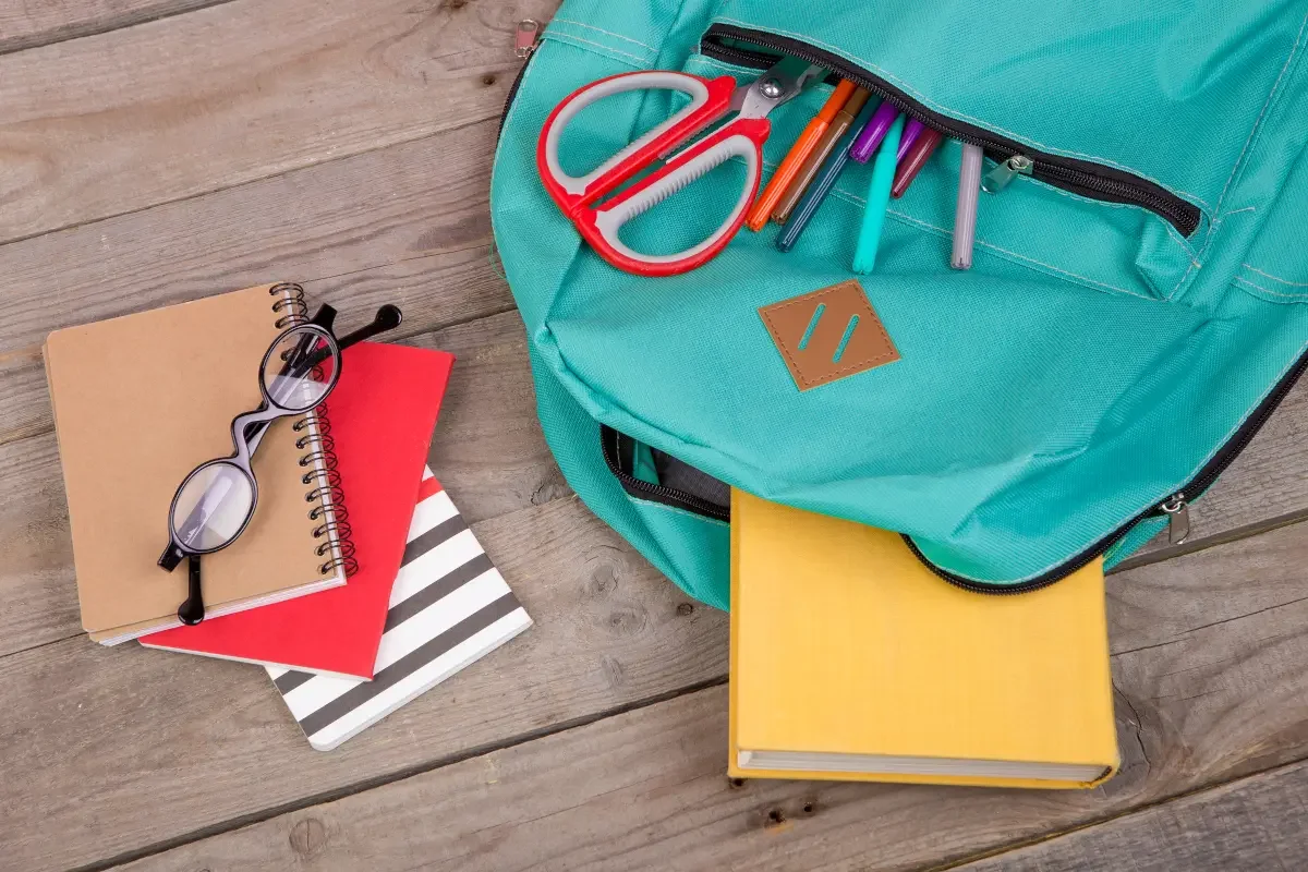 Open teal backpack with pens, scissors, and notebooks on a wooden table