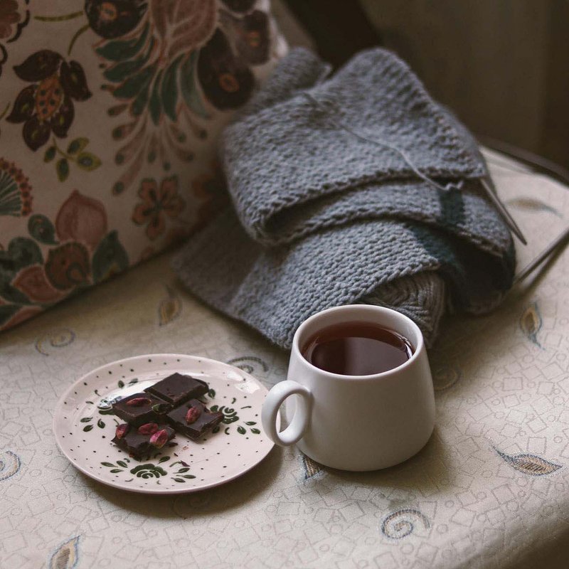 Cup of tea and chocolate on a cushion beside a knitted blanket